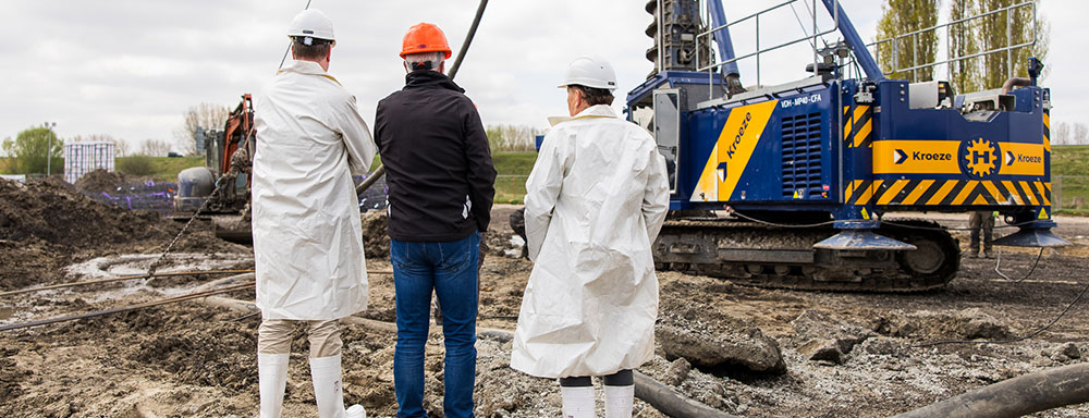 Three people, Jan van der Geest and Dirk Lorenz Meyer wearing white protective suits, observe a construction site where a pile-driving machine is in operation.