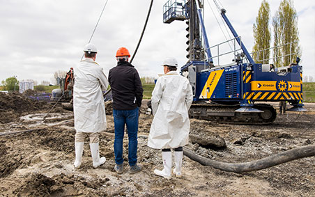Three people, Jan van der Geest and Dirk Lorenz Meyer wearing white protective suits, observe a construction site where a pile-driving machine is in operation.